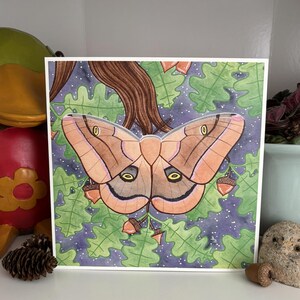 May include: Square art print featuring a large moth with tan and brown wings, set against a backdrop of green leaves, acorns, and a starry night sky. The moth's wings have intricate patterns. The artwork is framed in white.