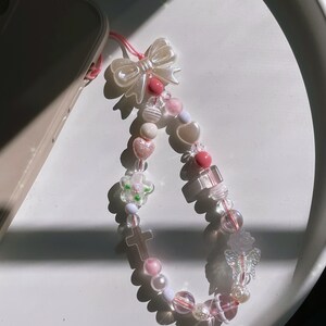 May include: A pink and white beaded phone charm with a pearl bow, cross, flower, and butterfly charms.