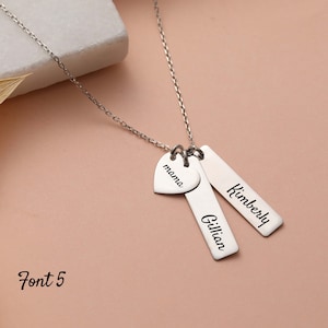 May include: Silver necklace with three charms: a heart charm with the word "mama" engraved on it, and two rectangular charms with the names "Kimberly" and "Gillian" engraved on them. The necklace is displayed on a light pink surface. Font 5.