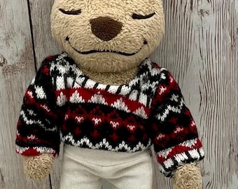 Può includere: Un orsacchiotto di peluche con un maglione fantasia e pantaloni bianchi. Il maglione presenta un motivo geometrico nero, rosso e bianco. L'orso ha gli occhi chiusi e un'espressione sorridente, suggerendo un'estetica amichevole e confortante.