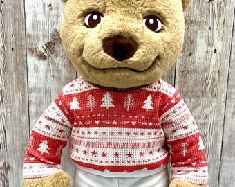 Può includere: Un orsacchiotto di peluche con un maglione natalizio rosso e bianco con un motivo ad albero e pantaloni bianchi. L'orso ha la pelliccia marrone, un naso marrone e un'espressione sorridente. Sono visibili le braccia e le gambe dell'orso.