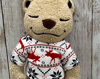 Può includere: Un orsacchiotto di peluche con gli occhi chiusi indossa un pigiama bianco con motivi rossi. Il pigiama presenta disegni di alci e fiocchi di neve. L'orso ha una pelliccia marrone chiaro e un naso marrone scuro.