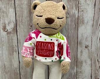 Può includere: Un orso di peluche marrone che indossa un maglione bianco e rosa con pois verdi e rossi e il testo "Seasons Greetings". L'orso indossa anche pantaloni bianchi.