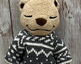 Può includere: Un orsacchiotto di peluche marrone che indossa un maglione di lana grigio e bianco con un motivo a fiocco di neve. L'orso è in piedi su uno sfondo di legno bianco.