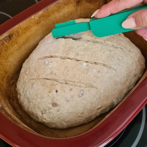 Op de afbeelding: Een brood met insnijdingen in de bovenkant, in een rode keramische bakvorm. Een groen plastic brood-insnijdingstool wordt gebruikt om de insnijdingen te maken.
