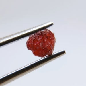 Puede incluir: Una piedra preciosa facetada de color rojo intenso sostenida por unas pinzas. La piedra preciosa tiene un aspecto áspero y natural.