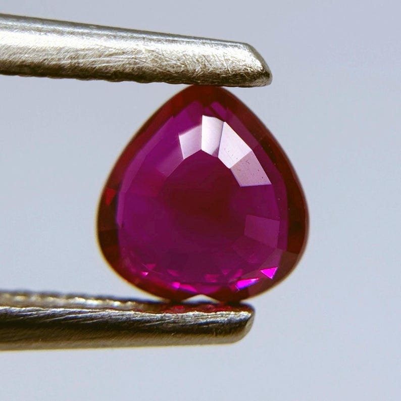 May include: A faceted, heart-shaped ruby gemstone held by metal tweezers. The ruby is a deep, vibrant red with multiple facets that reflect light. The tweezers are silver-colored, and the background is a soft, neutral tone.