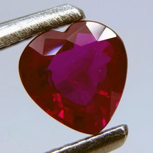 May include: A heart-shaped, faceted ruby gemstone. The ruby is a deep red color with a high degree of clarity and brilliance. The gemstone is held by a pair of silver tweezers, showcasing its shape and color.