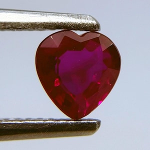 May include: A heart-shaped ruby gemstone, held by metal tweezers. The gemstone has a deep red color with a faceted surface, reflecting light. The gem's shape is a perfect heart, showcasing its polished facets.