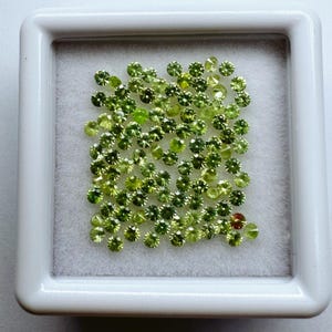 May include: A collection of small, round, light green gemstones displayed in a white, square container. The gemstones vary slightly in shade, with some appearing more yellow-green and others a deeper olive green. The container has a white, felt-like interior.
