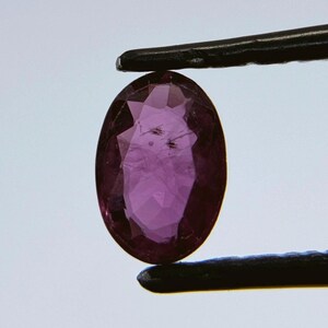 May include: A deep purple oval-shaped gemstone held by tweezers. The gemstone has a faceted cut and a shiny finish.