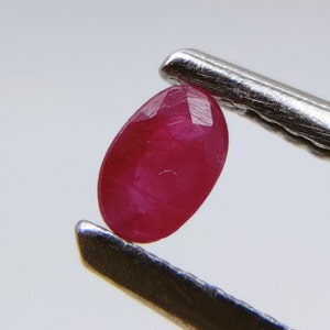 May include: A vibrant red oval-shaped gemstone, likely a ruby, held in a pair of tweezers.