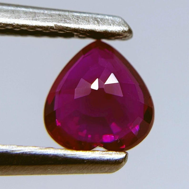 May include: A faceted, heart-shaped ruby gemstone held by a pair of silver tweezers. The ruby is a deep, rich red color with multiple facets that reflect light. The gemstone is approximately 0.5 inches in length.