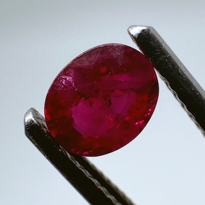 以下が含まれることがあります： 金属製のピンセットで保持された、鮮やかな赤色の楕円形の宝石。宝石は滑らかで磨かれた表面を持ち、深く豊かな色をしています。