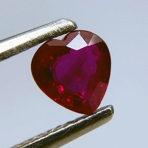 May include: A heart-shaped ruby gemstone, held by silver tweezers. The ruby has a deep red color with a vibrant, faceted surface. The gemstone is a symbol of love and passion.