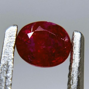 Puede incluir: Una piedra preciosa ovalada de color rojo oscuro sostenida por unas pinzas. La piedra preciosa tiene una superficie lisa y pulida y es transparente.