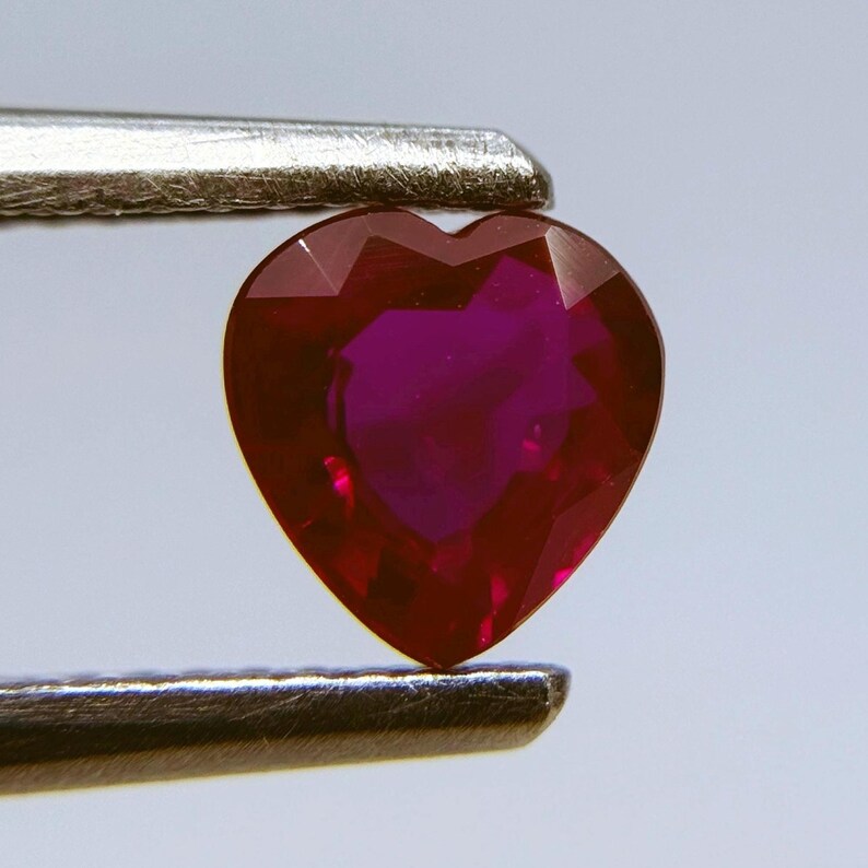 May include: A heart-shaped, faceted ruby gemstone held by a pair of tweezers. The ruby is a deep red color with a vibrant, purplish hue in the center. The gemstone's facets catch the light, creating a sparkling effect.