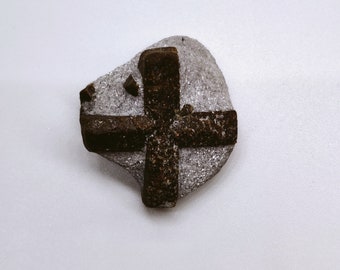 May include: A dark brown cross-shaped rock embedded in a light gray rock. The cross is made of multiple pieces of rock that have been joined together.