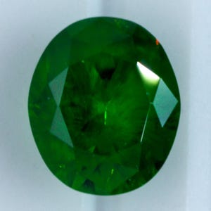 Puede incluir: Una gema de corte ovalado vibrante con un color verde esmeralda profundo y saturado. La piedra presenta una superficie facetada que crea reflejos geométricos nítidos y brillos, mostrando un alto nivel de claridad y un acabado pulido.