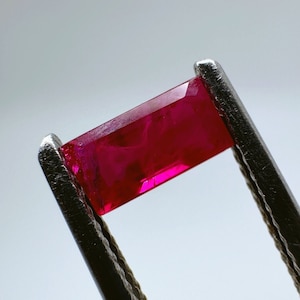 以下が含まれることがあります： 金属製のピンセットで保持された、長方形にカットされた鮮やかな赤い宝石。宝石は滑らかで磨かれており、わずかに輝いています。