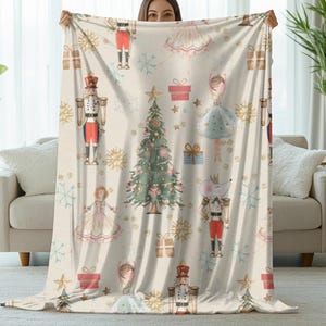 May include: A cream-colored blanket featuring a festive holiday design. The pattern includes illustrations of nutcrackers, ballerinas, Christmas trees, gift boxes, and snowflakes. The blanket is held up in front of a neutral background.