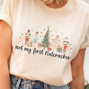 May include: Cream-colored t-shirt featuring a festive Nutcracker design with a Christmas tree, snowflakes, and characters. The text "not my first Nutcracker" is printed in black script. The shirt is paired with blue jeans.