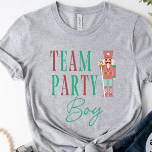 Może przedstawiać: Szara koszulka athletic heather z napisem "TEAM PARTY Boy" w kolorze czerwonym i zielonym oraz ilustracją postaci dziadka do orzechów. Koszulka jest zawiązana w pasie. Widoczne są również niebieskie dżinsy i okulary przeciwsłoneczne.