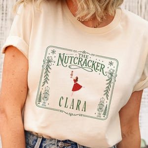 May include: A cream-colored t-shirt featuring a green rectangular frame with decorative bows and pine branches. Inside the frame, the text reads THE NUTCRACKER and CLARA, accompanied by a graphic of a dancer in a red dress and two nutcracker figures.