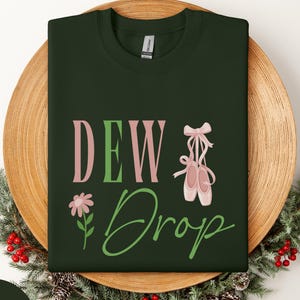 May include: Forest green sweatshirt with the text "DEW Drop" in pink and green, a flower, and ballet shoes. The sweatshirt is displayed on a wooden plate with a festive arrangement of greenery and red berries.