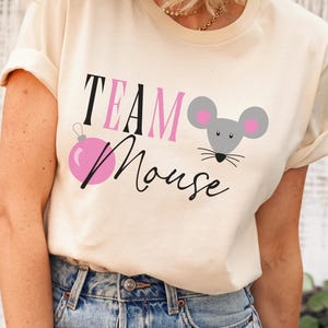 May include: Cream-colored t-shirt with the words "TEAM Mouse" in black and pink, along with a pink ornament and a gray mouse graphic with pink ears. The shirt has rolled-up sleeves.