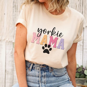 May include: A woman wearing a light beige t-shirt with the text "yorkie MAMA" in rainbow colors and a black paw print. She is wearing light blue jeans with distressed details.