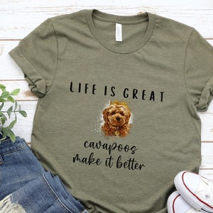 May include: Olive green t-shirt with the text "LIFE IS GREAT" above a watercolor image of a Cavapoo dog. Below the dog image, the text reads "cavapoos make it better". The shirt is paired with blue jeans and white sneakers.