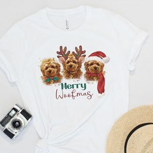 May include: White t-shirt with a Christmas-themed design featuring three brown dogs wearing festive attire. The dogs are wearing reindeer antlers, a Santa hat, and a red scarf. The text "Merry Woofmas" is printed below the dogs.