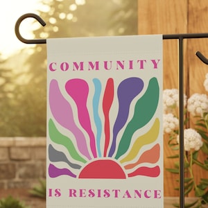 May include: A beige garden flag with a colorful sunrise design and the text "COMMUNITY IS RESISTANCE" in pink letters.