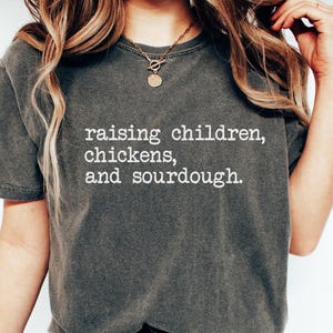 May include: A dark gray t-shirt with the text "raising children, chickens, and sourdough." printed in white.