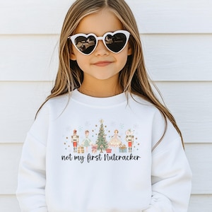 May include: A white long-sleeve sweatshirt featuring a graphic print of a Christmas tree, nutcrackers, and gift boxes with the text "not my first Nutcracker" in black script lettering across the center of the chest.
