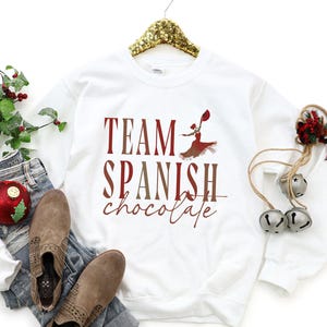 Pode incluir: Camisola branca com o texto "TEAM SPANISH chocolate" em bordô e castanho. Um gráfico de uma mulher em um vestido vermelho também está na camisa. A camisola é exibida com jeans e botas marrons.