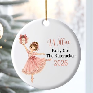 May include: A round white ceramic ornament featuring a brown-haired dancer in a pink tutu holding a gift box. The text reads Willow, Party Girl, The Nutcracker, and 2026. A gold string is attached to the top for hanging on a Christmas tree.