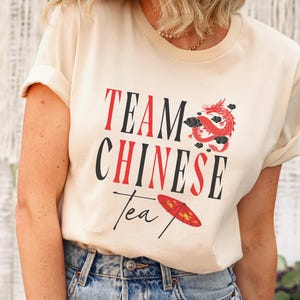 May include: Cream-colored t-shirt with the text "TEAM CHINESE Tea" in red and black. A red dragon and a red umbrella are featured in the design. The shirt is paired with blue jeans.