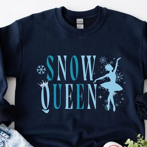 May include: Navy blue sweatshirt with the words "SNOW QUEEN" in light blue, along with a snowflake, crown, and ballerina graphic. The sweatshirt is folded on a light blue denim surface, with white sneakers and greenery visible.