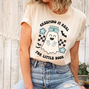 May include: Cream-colored t-shirt with the text "CREEPING IT REAL FOR LITTLE BOOS." The graphic features a smiling ghost wearing a nurse's cap, peace signs, bats, and flowers. The shirt is paired with blue jeans.