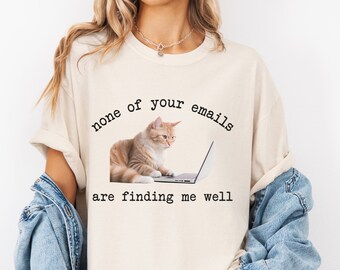 May include: A beige t-shirt with a graphic of a cat sitting on a laptop. The text on the shirt reads "none of your emails are finding me well."