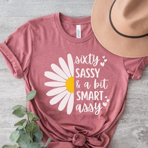 Pode incluir: T-shirt cor malva com o texto "sixty SASSY & a bit SMART assy" e um gráfico de margarida. Um chapéu bege repousa sobre a camisa. A camisa é amarrada na parte inferior. Ramos de eucalipto no canto inferior esquerdo.