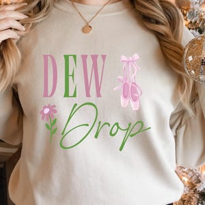 May include: Cream-colored sweatshirt with the text "DEW Drop" in pink and green. A pink flower and a pair of pink ballet shoes with a bow are also featured. The sweatshirt is a casual, comfortable style.