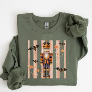 May include: A military green crewneck sweatshirt featuring a central graphic of a nutcracker dressed in orange and black, surrounded by flying bats and orange stars against a background of vertical light-colored stripes on the front chest.