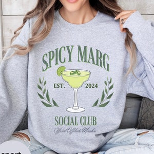 May include: A gray sweatshirt with the text "Spicy Marg Social Club" and a green illustration of a margarita glass with lime and jalapeños. The text "Est. 2024" is below the glass.
