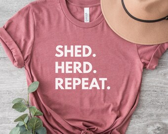 Puede incluir: Una camiseta rosa polvorienta con las palabras "SHED. HERD. REPEAT." en letras blancas. Un sombrero color canela con una banda negra descansa sobre la camisa. Se ve vegetación en la esquina inferior izquierda. La camisa está anudada en la cintura.