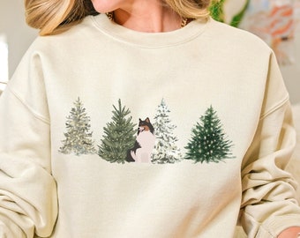 May include: A cream-colored sweatshirt with a graphic of a dog standing in front of four Christmas trees. The trees are decorated with lights and ornaments.