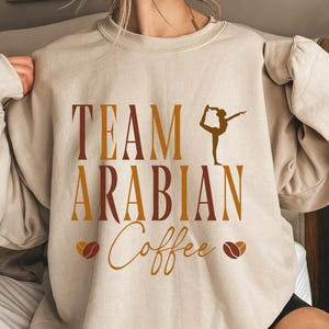 May include: Beige sweatshirt with the text "TEAM ARABIAN Coffee" in brown and gold lettering. A silhouette of a person in a yoga pose is above the word "Coffee", and coffee bean graphics are on either side of the word.