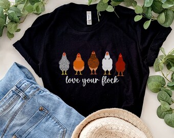 May include: Black t-shirt with a graphic of five chickens in a row and the text "love your flock". The shirt is laid out on a white surface with a pair of blue jeans and a straw hat.